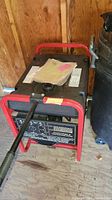 Full front view of portable generator showing control panel with brand and model label, red frame, black fuel tank, paper manuals on top.