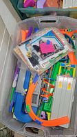 Plastic bin filled with Hot Wheels track pieces in orange and blue, with a plastic bag containing smaller items or pieces on top, also some paper items.