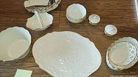Top view of off-white Lenox china including large oval platter, round scalloped dishes, lidded container and leaf-shaped dish with serving spoon laid out on a wood surface.