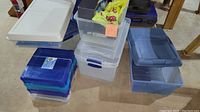 Overview of assorted storage totes stacked and placed on the floor, showing various sizes and colors including blue, clear, and white lids.
