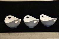 Three white ceramic bowls placed side by side against a dark background, each with a hole handle on the shorter ends, smooth finish, and modern design.