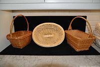 Three wicker baskets displayed on a black mat, two with handles and one oval shape without handle, placed on a marble surface.