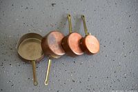 Four copper melting pots of varying sizes laid out showing copper exterior with patina and long metal handles.