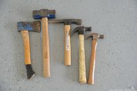 Top-down view of four hammers and one axe laid out on a concrete floor, showing variation in head styles and handle sizes.