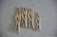 Four all metal locking pliers of various sizes laid out on a speckled floor surface. The pliers have different jaw shapes including straight, curved, and needle-nose.