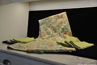 Photo showing the folded autumn-themed tablecloth in muted tones with leaf and floral patterns, accompanied by two stacks of cloth napkins in light and dark green shades.