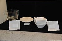 Photo showing one glass trifle bowl with a pedestal, a small white ceramic cake tray on pedestal with decorative pattern, and eight square white ceramic dessert bowls arranged in stacks.