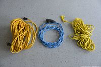 Three coiled cords and rope laid on the ground: a long yellow extension cord, a medium blue block heater cord, and a short thick yellow rope.