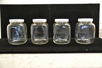 Front view of 4 clear glass jars with white plastic lids arranged in a row against a black background, showing jar size and lid shape clearly.