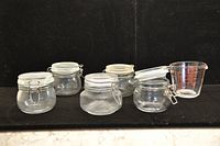Photo of 5 clear glass jars with bale wire locking lids arranged in a row on a black background with white marble surface beneath. Also included is a clear plastic measuring cup with red markings, positioned on the right end.