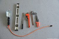 Photo showing a hanging work lamp with cage guard, spirit level, metal file, pipe wrench, carpenter's square, and tin snips spread on a gray floor