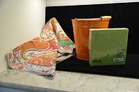 Photo showing an orange metal bucket, orange paisley patterned tablecloth, and sealed package of green IKEA napkins placed on a marble countertop against a white background.