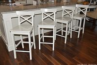 Four white IKEA kitchen stools arranged in a line in front of a kitchen island on dark hardwood floor. Each stool has a solid frame, backrest with X design, and footrests at 25-inch seat height.