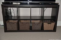 Full frontal view of black IKEA cubby storage unit with 8 sections, lower 4 containing woven baskets; unit beneath TV stand in carpeted room.