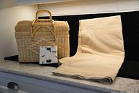 Photo showing closed wicker picnic basket, tan folded tablecloth, and package of ant-themed napkins on countertop.