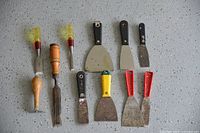 Ten spatulas and putty knives with wooden, plastic handles in various colors and metal blades, shown laid out on a light speckled surface.