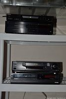 Four stereo components on metal shelving: Sony CD player and tape deck top shelf, Toshiba cassette deck and Nakamichi multi-disc CD player below.