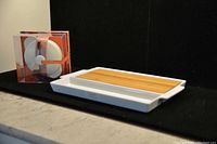 White ceramic cheese tray with removable wooden board and boxed Brie cheese baker set on marble counter with black backdrop.
