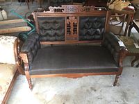 Full front view of antique settee displaying carved wood backrest, black tufted upholstery on back and arms, smooth seat fabric.