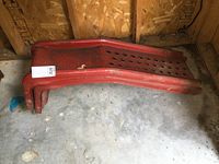 Pair of red steel automotive ramps stacked on concrete floor against wood wall, showing full length and surface holes for traction.