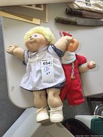 Pair of two 16-inch Cabbage Patch dolls lying on a table, one in white dress and the other in red jumpsuit holding bottle.
