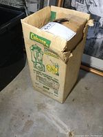 Cardboard box with Coleman branding and Sport-Lite lantern image on the side. Shows notable wear and tear on the box edges and top flaps.