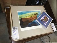 Three framed prints stacked on a small table showing the canoe print prominently with smaller prints on top.