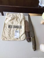 Lot showing hunting knife with sheath, brass-handled machete, and West Range Co. bag together on table.