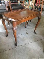 Front view of a wooden side table with curved legs and carved details, sitting on a concrete floor.