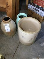 Three stoneware crocks on a concrete floor. Includes a large beige crock with markings, a plain tan/beige crock, and a pale green painted crock.