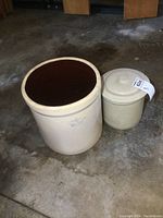 Photo showing both stoneware crocks together on concrete floor; one with lid and smaller, one open and larger.