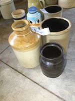 Group photo showing all three stoneware crocks together on a floor surface with various other items in background. Three crocks include one large tan and yellow, one medium beige, and one small dark brown.