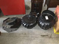 Pair of black metal turbine roof vents with circular bases and turbine tops, placed on concrete floor.