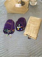 Three cloth bags on quilted surface: two purple velvet drawstring pouches with gold stitching and one beige cotton pouch, each containing assorted glass marbles.