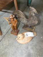 Three carved wooden figures including farmer with rake, rooster, and duck on concrete floor
