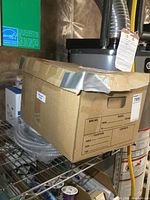 Closed cardboard box taped shut, labeled and placed on wire shelving.