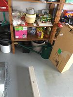 Overall view showing wooden shelving with gardening supplies, plastic and ceramic planters, white bucket and large green metal watering can on floor.