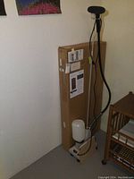 Full view of the Jiffy Steamer on wheels with attached steam wand and water reservoir near a cardboard box containing the Steamboard.