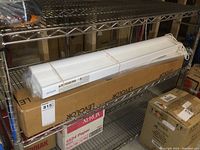 Three Levolor window blinds on metal shelf; two unboxed blinds bundled with elastic bands and one blind still inside sealed packaging showing brand name and size.