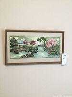 Framed needlepoint artwork depicting a garden landscape with bridge, pavilion, and flowering trees.