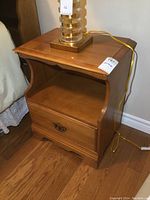 Front angle view of the solid maple Shermag nightstand showing drawer with decorative handle and open shelf space below the lamp on top.