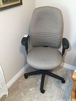 Front view of an ergonomic office chair with light gray fabric upholstery and black armrests on a five caster base.