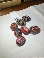 Seven hand-painted wooden eggs arranged on a white cloth, showcasing various floral and geometric patterns with different base colors like black and red.