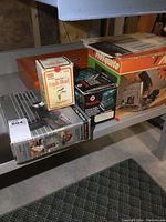 Photo shows multiple boxed power tools arranged on metal shelving in basement. The Bosch Mosquito jig saw and Craftsman angle head box are visible, along with a Makita sander box and a paint stripper box.