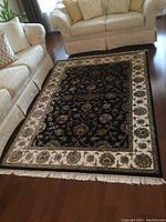 Full view of rectangular handwoven rug in living room setting, showing black field, cream border, and fringe ends