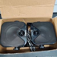 Two black water-resistant Herdio speakers with metal mesh grille and speaker wire inside brown cardboard box.
