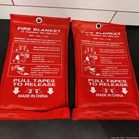 Two red fire blanket pouches with white text instructions and pull tapes visible, laying side by side on tiled surface