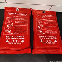 Two red pouch fire blankets side by side showing front with printed fire safety instructions and release tape pull areas. Size 1.0m x 1.0m noted.