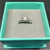 Close-up photo of a 2 carat round cut moissanite gem set in a smooth 925 sterling silver band ring, displayed inside a turquoise jewelry box with grey padded lining.