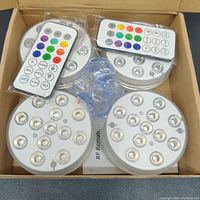 Four 3-inch round waterproof RGB LED lights in a box along with two remote controls in plastic bags, showing the lot contents all new and unused.